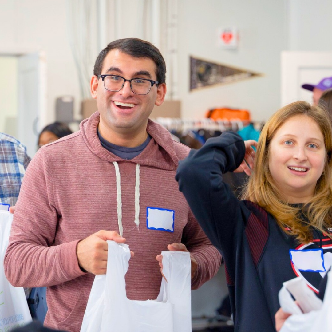 Two smiling volunteers wearing name tags hold white bags at an indoor service event, with other participants visible in the background.